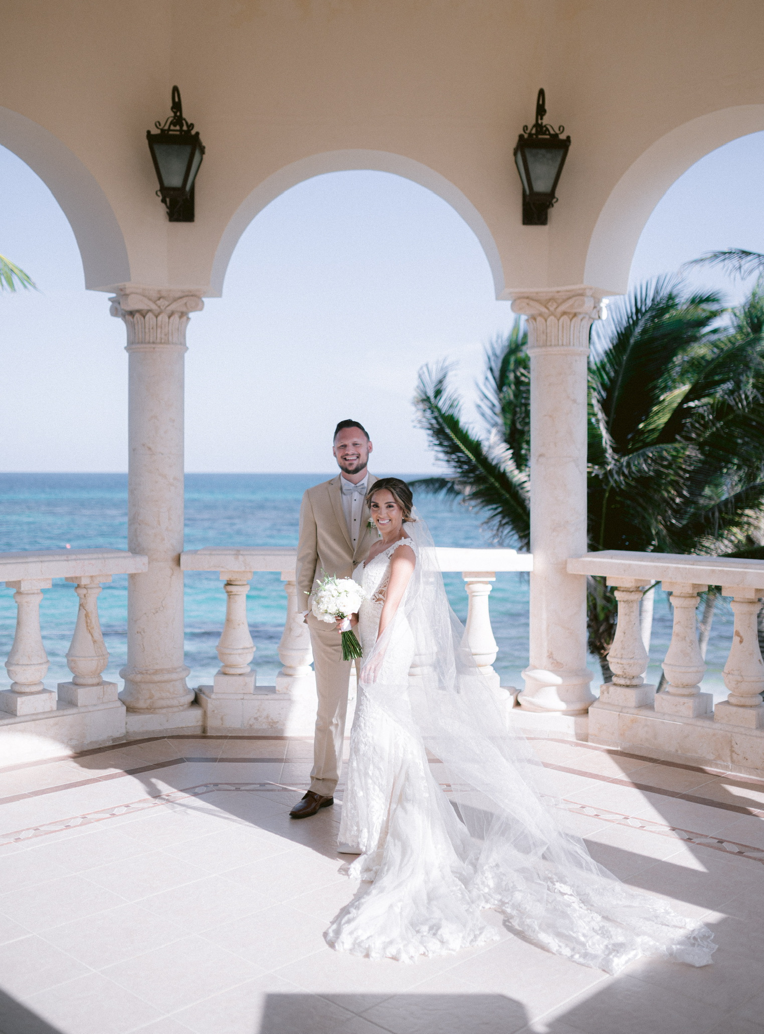 Joshua and Celeste Dendy together in wedding attire beneath seaside arches, with palm trees and the ocean behind them.